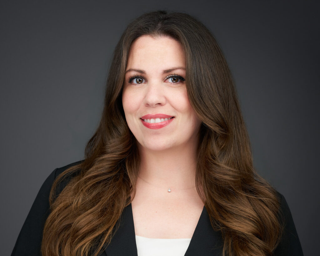 Close-up portrait of a smiling woman with long brown hair, wearing a black blazer and a delicate necklace, against a gray backdrop.